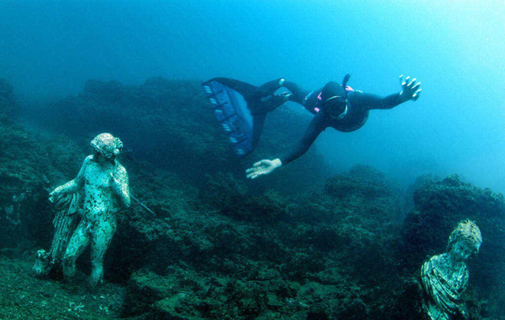 A diver swims between replica statues depicting Dionysus (left) and a girl (right) in Baiae.